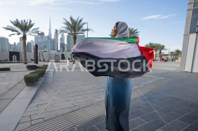 اليوم الوطني الإماراتي 2 ديسمبر،  الاحتفال بذكرى استقلال دولة الإمارات العربية المتحدة، امراة عربية خليجية اماراتية ترتدي العباءة والحجاب تضع علم الإمارات على كتفيها في النهار، التجول في الشوارع بايماءات السعادة والفخر والاعتزاز، مفهوم حب الوطن والانتماء اليه