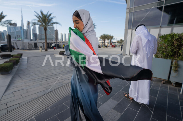 احياء اليوم الوطني الإماراتي 2 ديسمبر،  الاحتفال بذكرى استقلال دولة الإمارات العربية المتحدة، امراة عربية خليجية اماراتية ترتدي العباءة والحجاب تقبل علم الإمارات في النهار، التجول في الشوارع بايماءات السعادة والفخر والاعتزاز، مفهوم حب الوطن والانتماء اليه