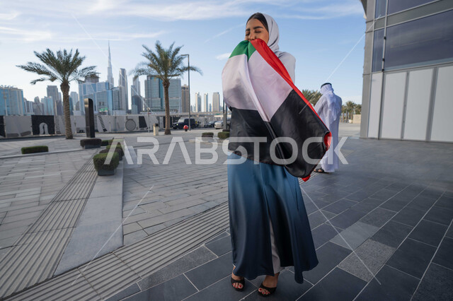 Celebrating the UAE National Day on December 2, celebrating the anniversary of the independence of the United Arab Emirates, an Emirati Gulf Arab woman wearing an abaya and hijab kissing the UAE flag during the day, walking in the streets with gestures of happiness, pride and honor, the concept of love of the homeland and belonging to it