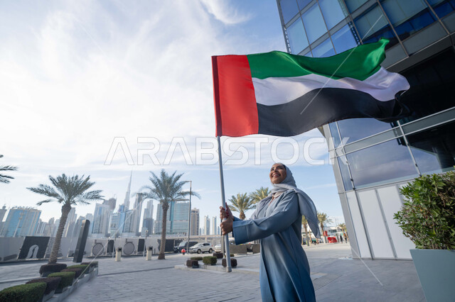 الاحتفال باليوم الوطني الإماراتي 2 ديسمبر، إحياء ذكرى استقلال دولة الإمارات العربية المتحدة، امراة عربية خليجية اماراتية ترتدي العباءة والحجاب ترفع علم الإمارات في النهار، التجول في الشوارع بايماءات السعادة والفخر والاعتزاز، مفهوم حب الوطن والانتماء اليه