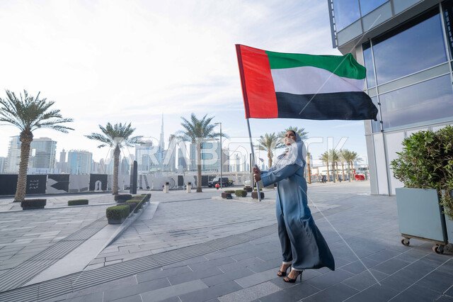 حب الوطن والانتماء اليه، الاحتفال باليوم الوطني الإماراتي 2 ديسمبر، إحياء ذكرى استقلال دولة الإمارات العربية المتحدة، امراة عربية خليجية اماراتية ترتدي العباءة والحجاب ترفع علم الإمارات في النهار، التجول في الشوارع بايماءات السعادة والفخر والاعتزاز