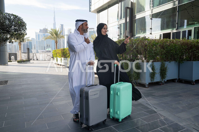 قضاء رحلة صيفية مسلية، مفهوم السياحة في إجازة الصيف، زوجان عربيان خليجيان اماراتيان يجران حقائب الامتعة امام الفندق، تعابير وايماءات السعادة والسرور، عائلة تقضي وقت ممتع، الاستعداد للسفر في جولة سياحية