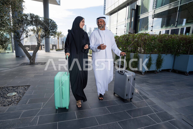Spending a fun summer trip, the concept of tourism during summer vacation, an Arab Gulf Emirati couple dragging their luggage in front of the hotel, expressions and gestures of happiness and pleasure, a family having a good time, preparing to travel on a tourist tour