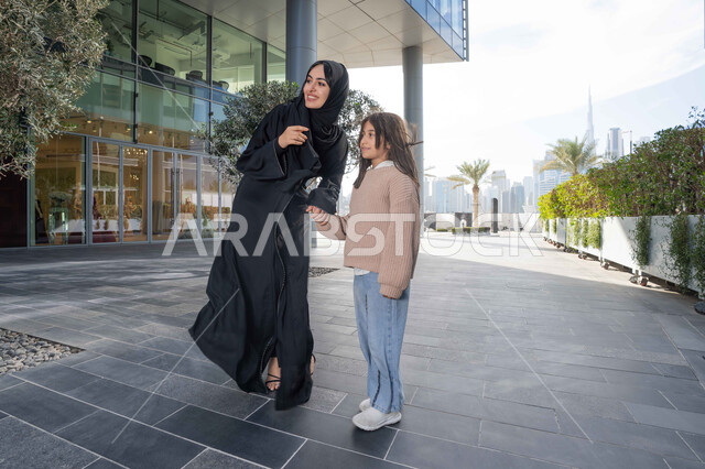 تبادل الأحاديث والأخبار بإيماءات السرور والفرح, أم عربية خليجية إماراتية ترتدي الحجاب والعباءة تقوم بالمشي والتجول في الهواء الطلق برفقة ابنتها، الاشارة الى شيء ما، عائلة سعودية تقوم بالتنزه في وقت العطلة في إحدى الاماكن العامة في الإمارات
