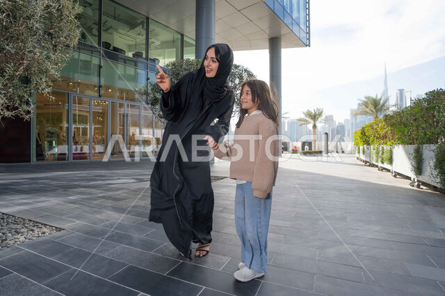 الاشارة الى شيء ما، عائلة سعودية تقوم بالتنزه في وقت العطلة في إحدى الاماكن العامة في الإمارات, تبادل الأحاديث والأخبار بإيماءات السرور والفرح, أم عربية خليجية إماراتية ترتدي الحجاب والعباءة تقوم بالمشي والتجول في الهواء الطلق برفقة ابنتها