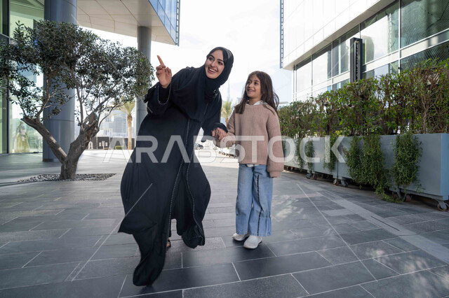 تبادل الأحاديث والأخبار بإيماءات السرور والفرح, أم عربية خليجية إماراتية ترتدي الحجاب والعباءة تقوم بالمشي والتجول في الهواء الطلق برفقة ابنتها، الاشارة الى شيء ما، عائلة سعودية تقوم بالتنزه في وقت العطلة في إحدى الاماكن العامة في الإمارات