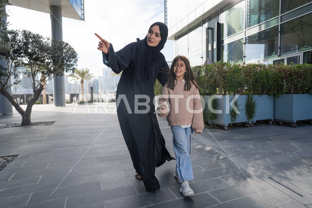 الاشارة الى شيء ما، عائلة سعودية تقوم بالتنزه في وقت العطلة في إحدى الاماكن العامة في الإمارات, تبادل الأحاديث والأخبار بإيماءات السرور والفرح, أم عربية خليجية إماراتية ترتدي الحجاب والعباءة تقوم بالمشي والتجول في الهواء الطلق برفقة ابنتها