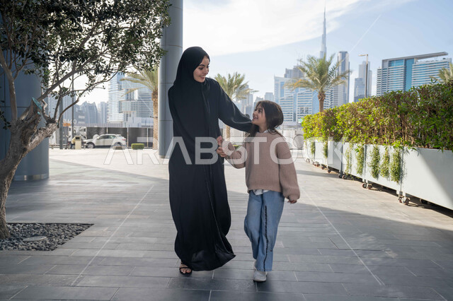 تبادل الأحاديث والأخبار بإيماءات السرور والفرح, أم عربية خليجية إماراتية ترتدي الحجاب والعباءة تقوم بالمشي والتجول في الهواء الطلق برفقة ابنتها، الاشارة الى شيء ما، عائلة سعودية تقوم بالتنزه في وقت العطلة في إحدى الاماكن العامة في الإمارات