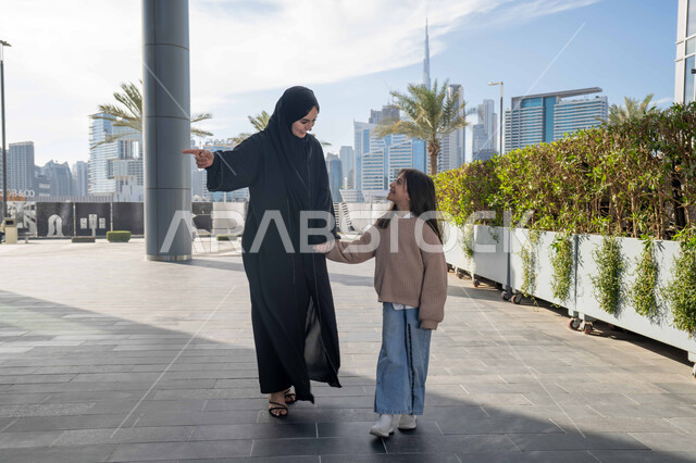 Pointing to something, a Saudi family taking a walk during the holiday in a public place in the Emirates, exchanging conversations and news with gestures of pleasure and joy, an Emirati Gulf Arab mother wearing a hijab and abaya walking and strolling in the open air with her daughter