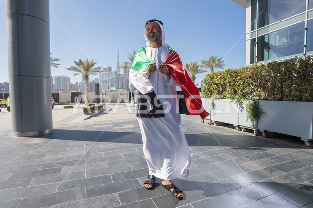 الاحتفال بذكرى تأسيس الاتحاد 2 ديسمبر في الطريق، التجول في الهواء الطلق، يوم العلم 3 نوفمبر، حب الوطن والاعتزاز به، اليوم الوطني الاماراتي 1971، رجل عربي خليجي اماراتي يرتدي الكندورة والغترة التقليدية يضع علم الامارات العربية المتحدة على كتفيه في النهار