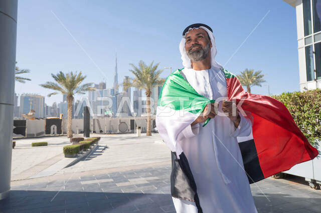 الاحتفال بذكرى تأسيس الاتحاد 2 ديسمبر في الطريق، التجول في الهواء الطلق، يوم العلم 3 نوفمبر، حب الوطن والاعتزاز به، اليوم الوطني الاماراتي 1971، رجل عربي خليجي اماراتي يرتدي الكندورة والغترة التقليدية يضع علم الامارات العربية المتحدة على كتفيه في النهار