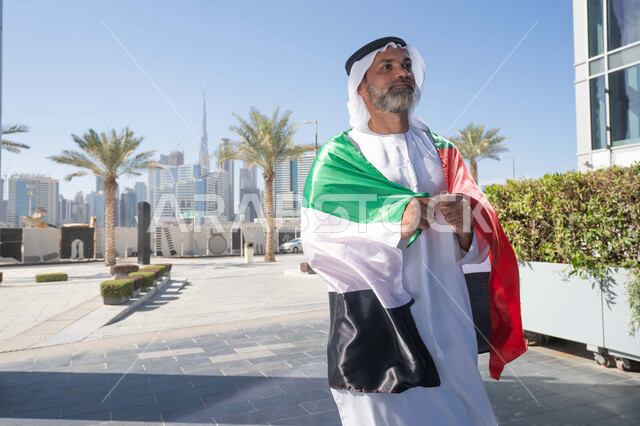 الاحتفال بذكرى تأسيس الاتحاد 2 ديسمبر في الطريق، التجول في الهواء الطلق، يوم العلم 3 نوفمبر، حب الوطن والاعتزاز به، اليوم الوطني الاماراتي 1971، رجل عربي خليجي اماراتي يرتدي الكندورة والغترة التقليدية يضع علم الامارات العربية المتحدة على كتفيه في النهار