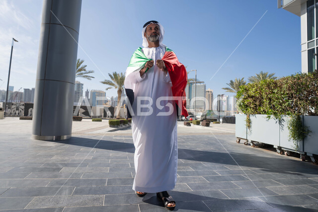Celebrating the anniversary of the founding of the Union, December 2, on the road, outdoor walk, Flag Day, November 3, love and pride of the homeland, UAE National Day 1971, an Arab Gulf Emirati man wearing the traditional ghutra kandara, placing the UAE flag on his shoulders during the day