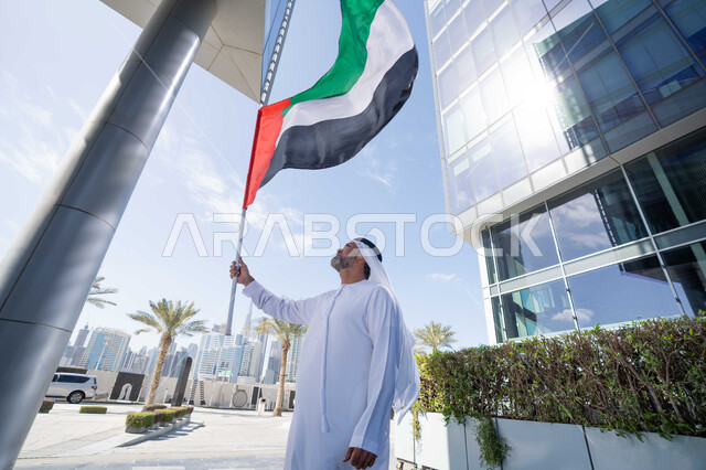 رفع راية البلاد للاعلى، الاحتفال بذكرى تأسيس الاتحاد 2 ديسمبر في الطريق، التجول في الهواء الطلق، يوم العلم 3 نوفمبر، حب الوطن والاعتزاز به، اليوم الوطني الاماراتي 1971، رجل عربي خليجي اماراتي يرتدي الكندورة والغترة يمسك علم الامارات العربية المتحدة في النهار