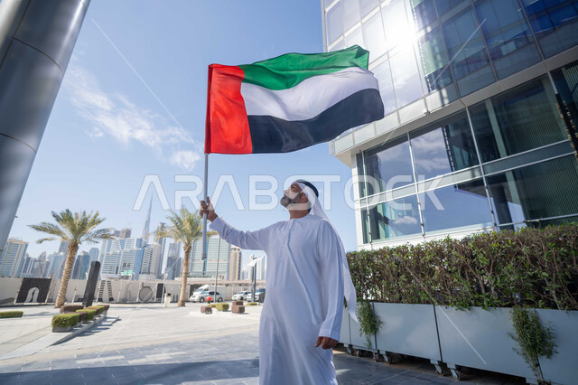 رفع راية البلاد للاعلى، الاحتفال بذكرى تأسيس الاتحاد 2 ديسمبر في الطريق، التجول في الهواء الطلق، يوم العلم 3 نوفمبر، حب الوطن والاعتزاز به، اليوم الوطني الاماراتي 1971، رجل عربي خليجي اماراتي يرتدي الكندورة والغترة يمسك علم الامارات العربية المتحدة في النهار