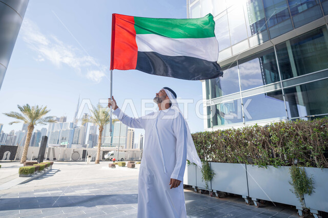 حب الوطن والاعتزاز به، اليوم الوطني الاماراتي 1971، رجل عربي خليجي اماراتي يرتدي الكندورة والغترة يمسك علم الامارات العربية المتحدة في النهار، رفع راية البلاد للاعلى، الاحتفال بذكرى تأسيس الاتحاد 2 ديسمبر في الطريق، التجول في الهواء الطلق، يوم العلم 3 نوفمبر