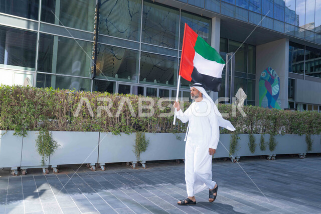 رفع راية البلاد للاعلى، الاحتفال بذكرى تأسيس الاتحاد 2 ديسمبر في الطريق، التجول في الهواء الطلق، يوم العلم 3 نوفمبر، حب الوطن والاعتزاز به، اليوم الوطني الاماراتي 1971، رجل عربي خليجي اماراتي يرتدي الكندورة والغترة يمسك علم الامارات العربية المتحدة في النهار