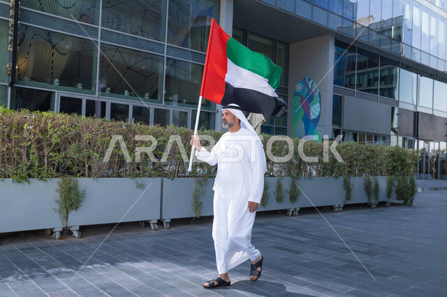 رفع راية البلاد للاعلى، الاحتفال بذكرى تأسيس الاتحاد 2 ديسمبر في الطريق، التجول في الهواء الطلق، يوم العلم 3 نوفمبر، حب الوطن والاعتزاز به، اليوم الوطني الاماراتي 1971، رجل عربي خليجي اماراتي يرتدي الكندورة والغترة يمسك علم الامارات العربية المتحدة في النهار