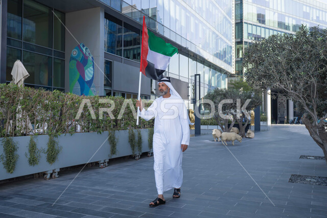 حب الوطن والاعتزاز به، اليوم الوطني الاماراتي 1971، رجل عربي خليجي اماراتي يرتدي الكندورة والغترة يمسك علم الامارات العربية المتحدة في النهار، رفع راية البلاد للاعلى، الاحتفال بذكرى تأسيس الاتحاد 2 ديسمبر في الطريق، التجول في الهواء الطلق، يوم العلم 3 نوفمبر
