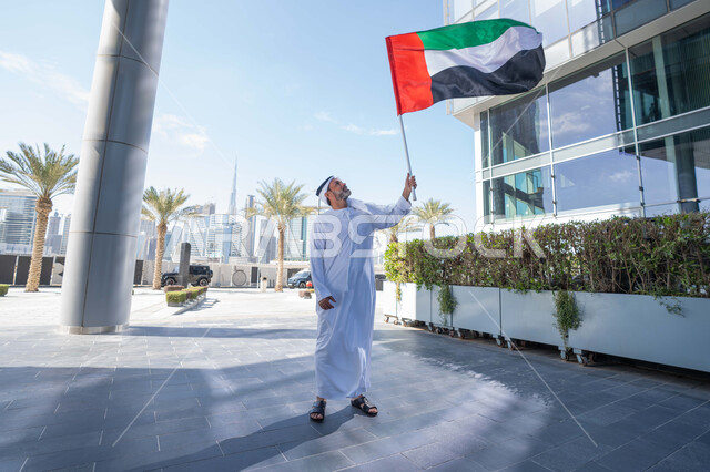 حب الوطن والاعتزاز به، اليوم الوطني الاماراتي 1971، رجل عربي خليجي اماراتي يرتدي الكندورة والغترة يمسك علم الامارات العربية المتحدة في النهار، رفع راية البلاد للاعلى، الاحتفال بذكرى تأسيس الاتحاد 2 ديسمبر في الطريق، التجول في الهواء الطلق، يوم العلم 3 نوفمبر