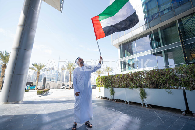 رفع راية البلاد للاعلى، الاحتفال بذكرى تأسيس الاتحاد 2 ديسمبر في الطريق، التجول في الهواء الطلق، يوم العلم 3 نوفمبر، حب الوطن والاعتزاز به، اليوم الوطني الاماراتي 1971، رجل عربي خليجي اماراتي يرتدي الكندورة والغترة يمسك علم الامارات العربية المتحدة في النهار