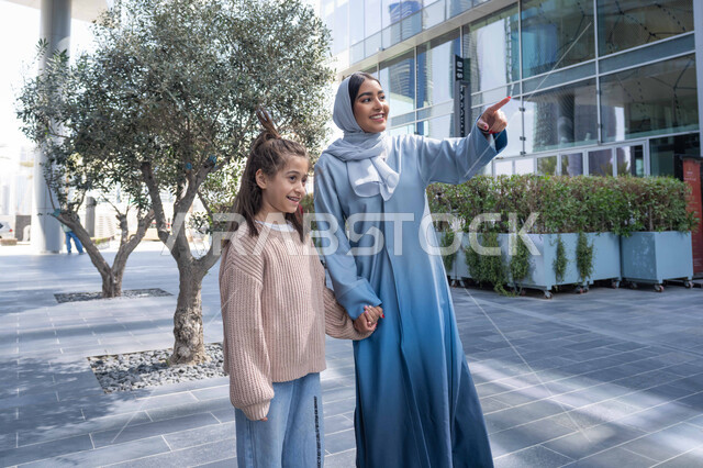 الاشارة الى شيء ما، عائلة سعودية تقوم بالتنزه في وقت العطلة في إحدى الاماكن العامة في الإمارات, تبادل الأحاديث والأخبار بإيماءات السرور والفرح, أم عربية خليجية إماراتية ترتدي الحجاب والعباءة تقوم بالمشي والتجول في الهواء الطلق برفقة ابنتها