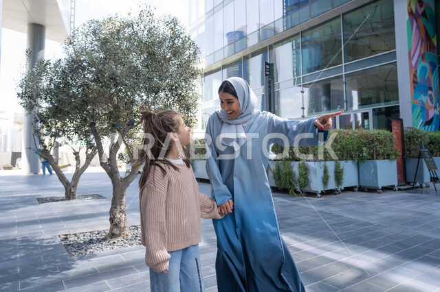 تبادل الأحاديث والأخبار بإيماءات السرور والفرح, أم عربية خليجية إماراتية ترتدي الحجاب والعباءة تقوم بالمشي والتجول في الهواء الطلق برفقة ابنتها، الاشارة الى شيء ما، عائلة سعودية تقوم بالتنزه في وقت العطلة في إحدى الاماكن العامة في الإمارات