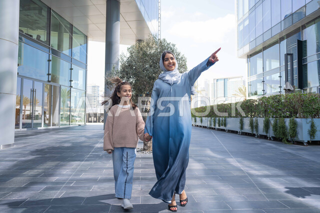 الاشارة الى شيء ما، عائلة سعودية تقوم بالتنزه في وقت العطلة في إحدى الاماكن العامة في الإمارات, تبادل الأحاديث والأخبار بإيماءات السرور والفرح, أم عربية خليجية إماراتية ترتدي الحجاب والعباءة تقوم بالمشي والتجول في الهواء الطلق برفقة ابنتها
