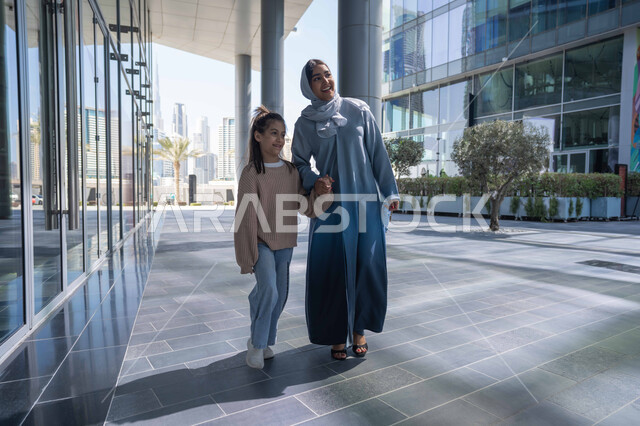 تبادل الأحاديث والأخبار بإيماءات السرور والفرح, أم عربية خليجية إماراتية ترتدي الحجاب والعباءة تقوم بالمشي والتجول في الهواء الطلق برفقة ابنتها، الاشارة الى شيء ما، عائلة سعودية تقوم بالتنزه في وقت العطلة في إحدى الاماكن العامة في الإمارات