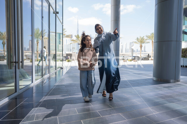 الاشارة الى شيء ما، عائلة سعودية تقوم بالتنزه في وقت العطلة في إحدى الاماكن العامة في الإمارات, تبادل الأحاديث والأخبار بإيماءات السرور والفرح, أم عربية خليجية إماراتية ترتدي الحجاب والعباءة تقوم بالمشي والتجول في الهواء الطلق برفقة ابنتها