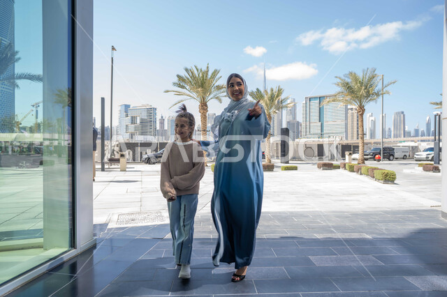 عائلة سعودية تقوم بالتنزه في وقت العطلة في إحدى الاماكن العامة في الإمارات, تبادل الأحاديث والأخبار بإيماءات السرور والفرح, أم عربية خليجية إماراتية ترتدي الحجاب والعباءة تقوم بالمشي والتجول في الهواء الطلق برفقة ابنتها، الاشارة الى شيء ما