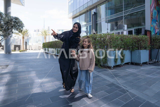 Pointing to something, a Saudi family taking a walk during the holiday in a public place in the Emirates, exchanging conversations and news with gestures of pleasure and joy, an Emirati Gulf Arab mother wearing a hijab and abaya walking and strolling in the open air with her daughter