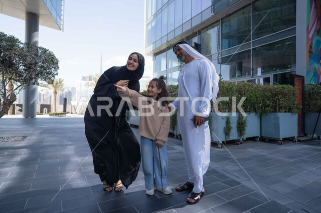 التنزه في وقت العطلة في إحدى الاماكن العامة في الإمارات, تبادل الأحاديث والأخبار بإيماءات السرور والفرح, زوجان عربيان خليجيان إماراتيان يقومان بالمشي والتجول في الهواء الطلق برفقة ابنتهم, الذكريات واللحظات العائلية السعيدة, الاشارة الى شيء ما