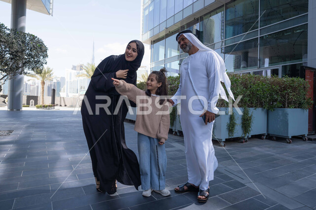 A walk during the holiday in a public place in the Emirates, exchanging conversations and news with gestures of pleasure and joy, an Emirati Gulf Arab couple walking and strolling in the open air with their daughter, happy family memories and moments, pointing to something