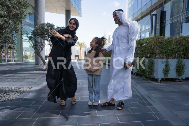 التنزه في وقت العطلة في إحدى الاماكن العامة في الإمارات, تبادل الأحاديث والأخبار بإيماءات السرور والفرح, زوجان عربيان خليجيان إماراتيان يقومان بالمشي والتجول في الهواء الطلق برفقة ابنتهم, الذكريات واللحظات العائلية السعيدة, الاشارة الى شيء ما