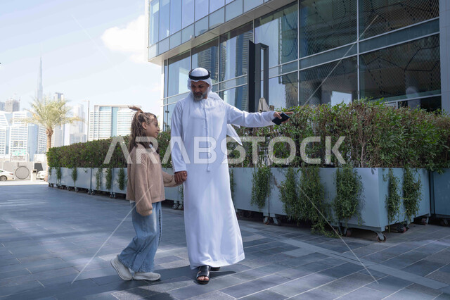 الاشارة الى شيء ما، التنزه في وقت العطلة في إحدى الاماكن العامة في الإمارات, تبادل الأحاديث والأخبار بإيماءات السرور والفرح, أب عربي خليجي إماراتي يرتدي الكندورة والغترة يقوم بالمشي والتجول في الهواء الطلق برفقة ابنته, الذكريات واللحظات العائلية السعيدة