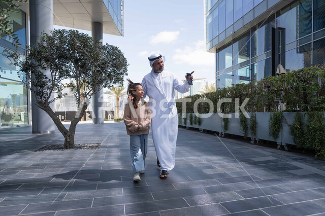 الاشارة الى شيء ما، التنزه في وقت العطلة في إحدى الاماكن العامة في الإمارات, تبادل الأحاديث والأخبار بإيماءات السرور والفرح, أب عربي خليجي إماراتي يرتدي الكندورة والغترة يقوم بالمشي والتجول في الهواء الطلق برفقة ابنته, الذكريات واللحظات العائلية السعيدة