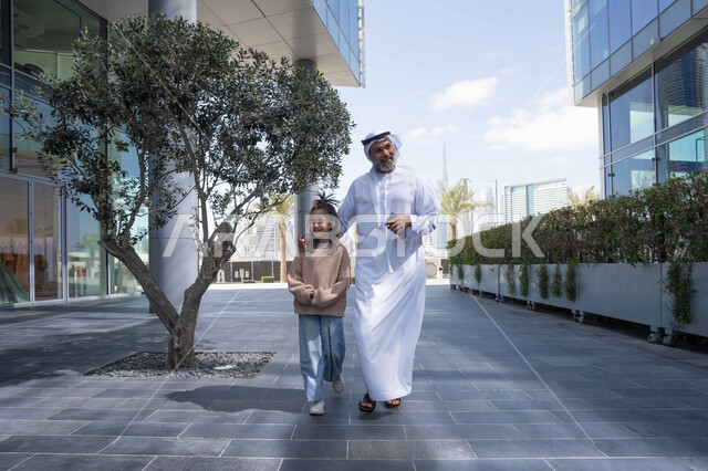 تبادل الأحاديث والأخبار بإيماءات السرور والفرح, أب عربي خليجي إماراتي يرتدي الكندورة والغترة يقوم بالمشي والتجول في الهواء الطلق برفقة ابنته, الذكريات واللحظات العائلية السعيدة، التنزه في وقت العطلة في إحدى الاماكن العامة في الإمارات