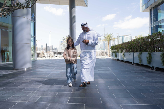 تبادل الأحاديث والأخبار بإيماءات السرور والفرح, أب عربي خليجي إماراتي يرتدي الكندورة والغترة يقوم بالمشي والتجول في الهواء الطلق برفقة ابنته, الذكريات واللحظات العائلية السعيدة، التنزه في وقت العطلة في إحدى الاماكن العامة في الإمارات