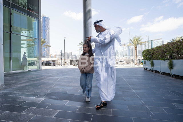 الاشارة الى شيء ما، التنزه في وقت العطلة في إحدى الاماكن العامة في الإمارات, تبادل الأحاديث والأخبار بإيماءات السرور والفرح, أب عربي خليجي إماراتي يرتدي الكندورة والغترة يقوم بالمشي والتجول في الهواء الطلق برفقة ابنته, الذكريات واللحظات العائلية السعيدة