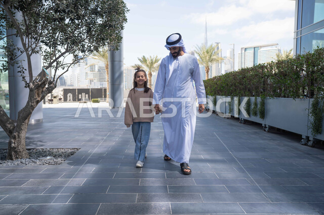 تبادل الأحاديث والأخبار بإيماءات السرور والفرح, أب عربي خليجي إماراتي يرتدي الكندورة والغترة يقوم بالمشي والتجول في الهواء الطلق برفقة ابنته, الذكريات واللحظات العائلية السعيدة، التنزه في وقت العطلة في إحدى الاماكن العامة في الإمارات