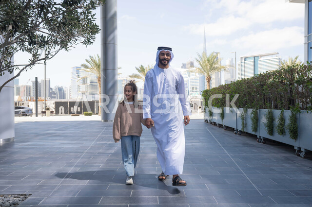 تبادل الأحاديث والأخبار بإيماءات السرور والفرح, أب عربي خليجي إماراتي يرتدي الكندورة والغترة يقوم بالمشي والتجول في الهواء الطلق برفقة ابنته, الذكريات واللحظات العائلية السعيدة، التنزه في وقت العطلة في إحدى الاماكن العامة في الإمارات