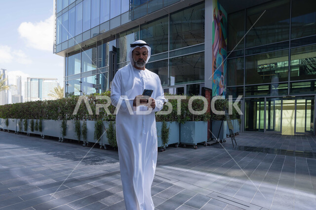 استخدام جهاز تقني حديث متطور في الحياة اليومية، مفهوم الاناقة والرجولة والاهتمام بالمظهر الخارجي، رجل عربي خليجي اماراتي يرتدي الغترة والكندورة التقليدية يمسك بيده الهاتف المحمول، خلفية مبنى احدى الشركات في الامارات العربية المتحدة، التجول في الهواء الطلق