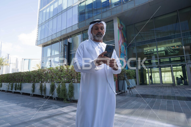 Using a modern, advanced technical device in daily life, the concept of elegance, masculinity, and interest in one's outward appearance, an Arab Gulf Emirati man wearing a traditional ghutra and kandura holding a mobile phone, background of a company building in the United Arab Emirates, walking outdoors