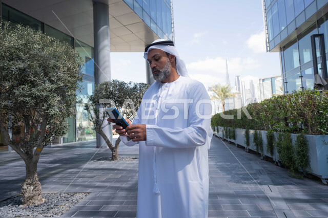 Using a modern, advanced technical device in daily life, the concept of elegance, masculinity, and interest in one's outward appearance, an Arab Gulf Emirati man wearing a traditional ghutra and kandura holding a mobile phone, background of a company building in the United Arab Emirates, walking outdoors
