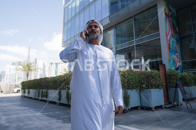 اجراء مكالمة هاتفية، استخدام جهاز تقني حديث متطور في الحياة اليومية، الاناقة والاهتمام بالمظهر الخارجي، رجل عربي خليجي اماراتي يرتدي الغترة والكندورة التقليدية يقوم بالرد على الهاتف المحمول امام مبنى احدى الشركات في الامارات العربية المتحدة، التجول في الهواء الطلق