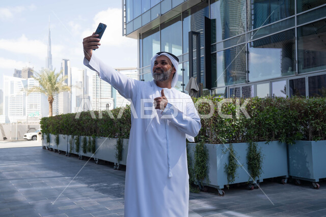 Taking selfies, using a modern, advanced technical device in daily life, elegance and attention to one's appearance, an Emirati Gulf Arab man wearing a traditional ghutra and kandura holding a mobile phone in front of a company building in the United Arab Emirates, walking outdoors
