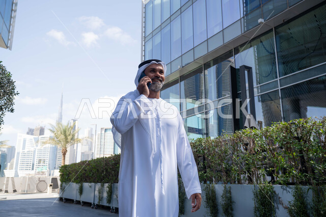 اجراء مكالمة هاتفية، استخدام جهاز تقني حديث متطور في الحياة اليومية، الاناقة والاهتمام بالمظهر الخارجي، رجل عربي خليجي اماراتي يرتدي الغترة والكندورة التقليدية يقوم بالرد على الهاتف المحمول امام مبنى احدى الشركات في الامارات العربية المتحدة، التجول في الهواء الطلق