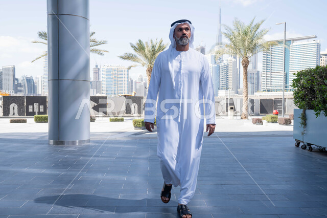 انتهاء وقت الدوام الرسمي، رجل عربي خليجي اماراتي يرتدي الغترة والكندورة التقليدية يخرج من الشركة بايماءات الثقة بالنفس، الحضور المبكر والنشاط الصباحي للعمل، بيئة العمل المريحة في شركات الامارات العربية المتحدة، التجول في الهواء الطلق