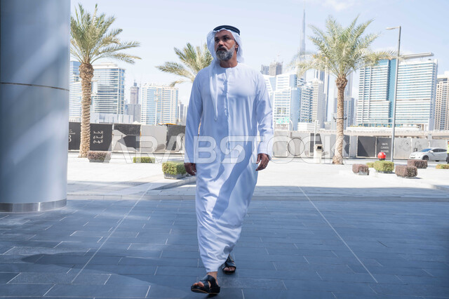 بيئة العمل المريحة في شركات الامارات العربية المتحدة، التجول في الهواء الطلق، انتهاء وقت الدوام الرسمي، رجل عربي خليجي اماراتي يرتدي الغترة والكندورة التقليدية يخرج من الشركة بايماءات الثقة بالنفس، الحضور المبكر والنشاط الصباحي للعمل
